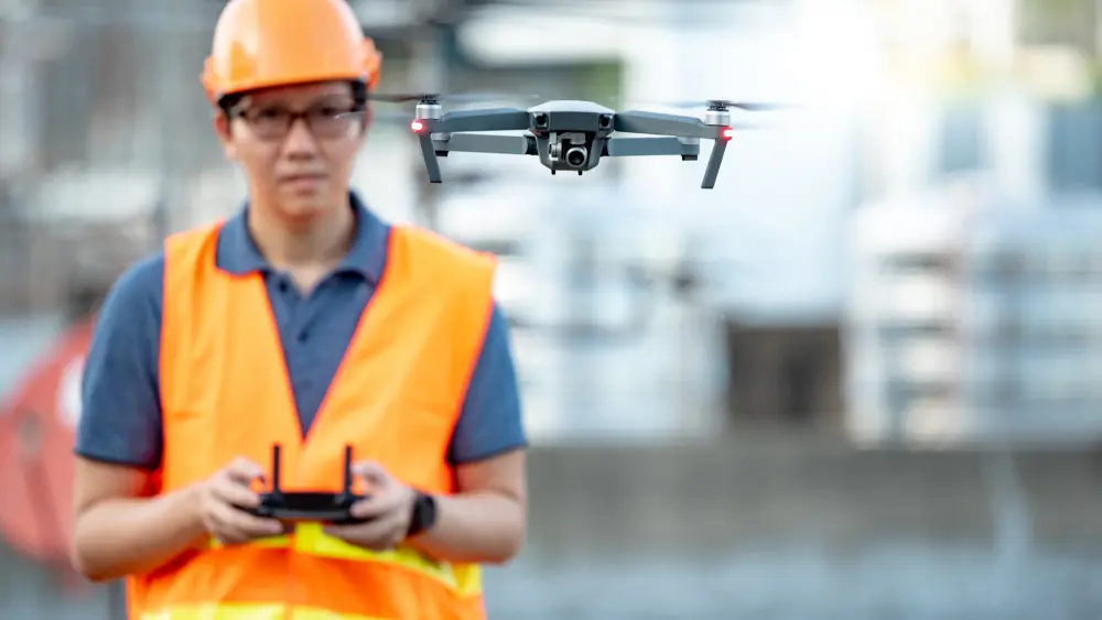 Surveyor in a hard hat and high-visibility vest flying a camera drone with a remote controller