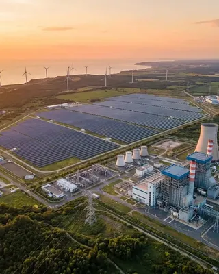 Coastal power generation site at sunset combining a solar array, wind turbines, cooling towers, a gas plant and a high-voltage switchyard