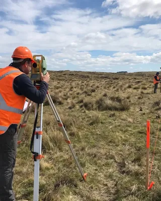 Survey crew running a total station and GNSS rover over open ground, with marker stakes set along the traverse